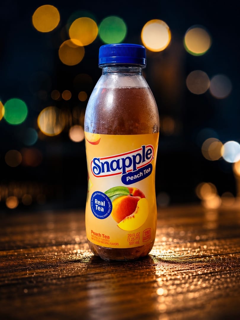 A Snapple Peach Tea bottle with a blue cap sits on a wooden surface with bokeh lights.