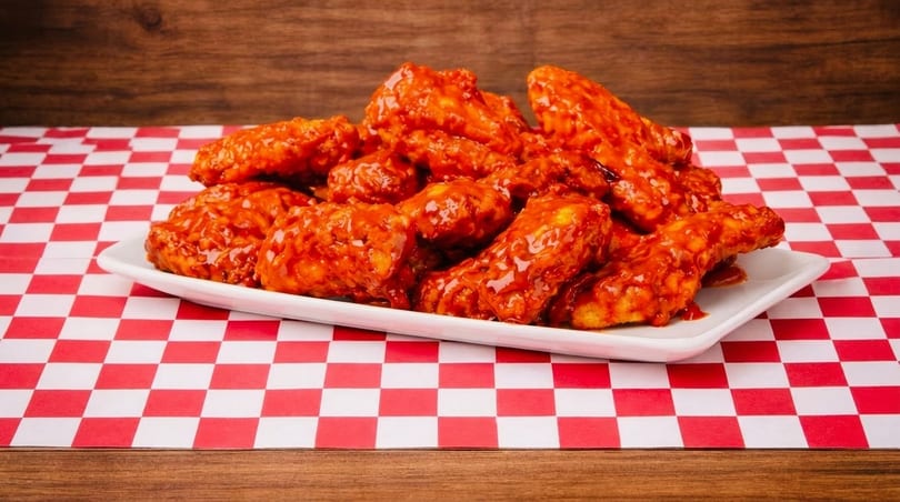 A white platter overflowing with glistening, saucy buffalo wings on a red and white checkered tablecloth.