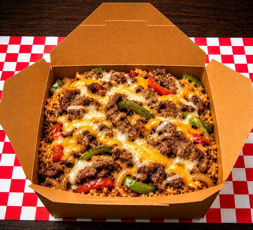 Cheesy ground beef and rice with peppers and onions in a brown takeout box on a red and white checkered tablecloth.