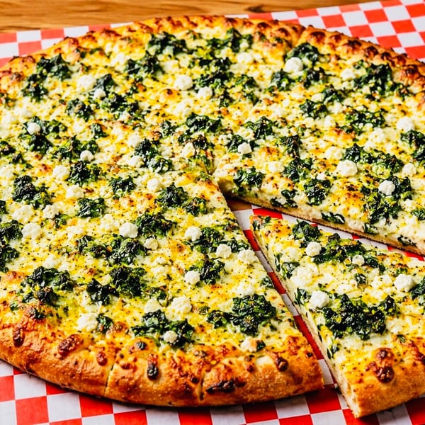 A whole spinach and feta pizza with one slice removed, on a red and white checkered paper.