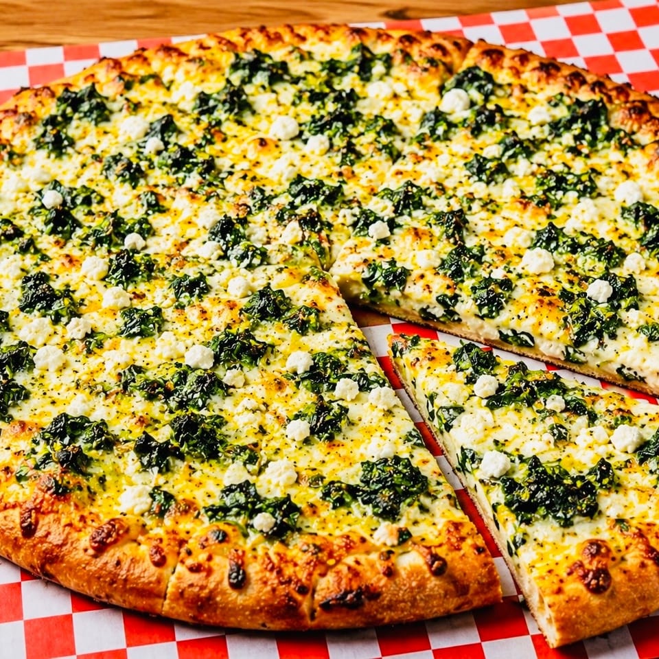 A whole spinach and feta pizza with one slice removed, on a red and white checkered paper.