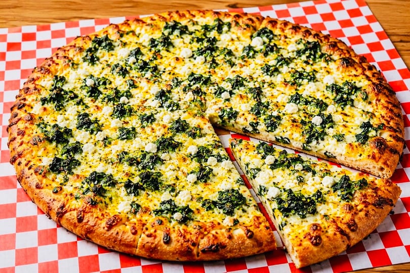 A whole spinach and feta pizza with one slice removed, on a red and white checkered paper.