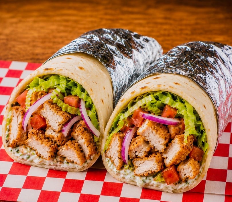Two chicken wraps filled with crispy chicken, lettuce, tomatoes, and red onions on a checkered paper.