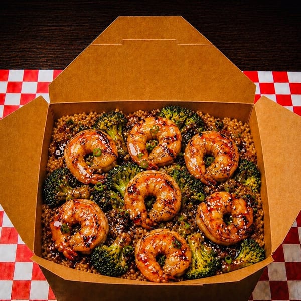 Takeout box filled with grilled shrimp, broccoli, and quinoa on a red and white checkered tablecloth.