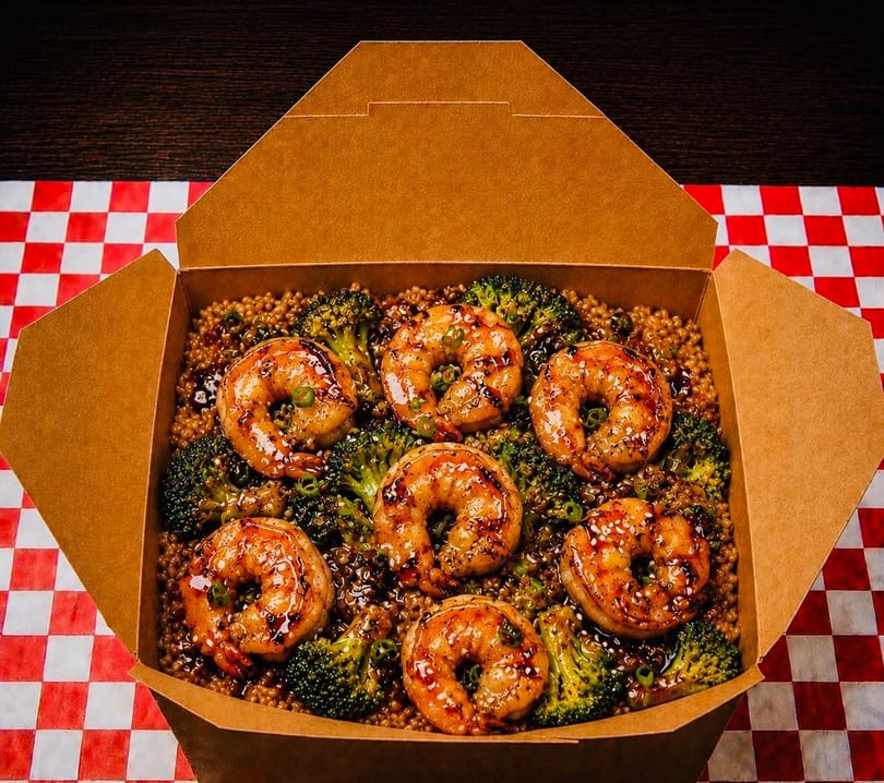 Takeout box filled with grilled shrimp, broccoli, and quinoa on a red and white checkered tablecloth.