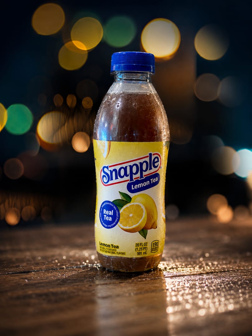 A cold Snapple Lemon Tea bottle with condensation sits on a wet wooden surface, with bokeh lights in the background.