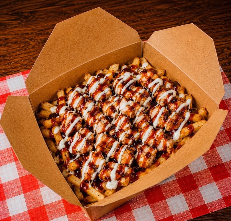 Loaded fries with grilled chicken, BBQ sauce, and white sauce in a cardboard box on a red and white checkered tablecloth.