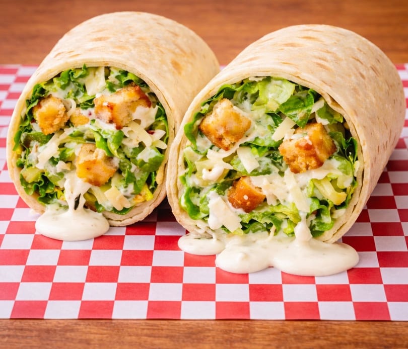 Two Caesar salad wraps, filled with lettuce, croutons, cheese, and dressing, on red and white checkered paper.