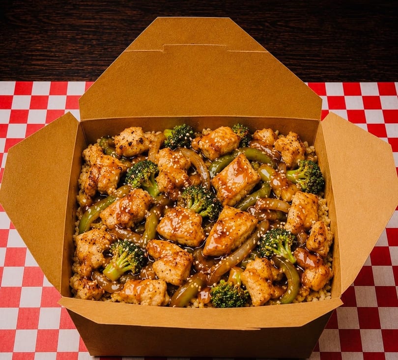 Chicken and broccoli stir-fry with sesame seeds in a brown takeout box on a red and white checkered tablecloth.