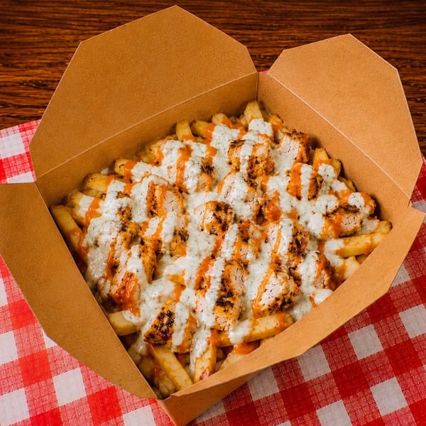 Fries topped with grilled chicken pieces, white sauce, and orange sauce in a cardboard box.