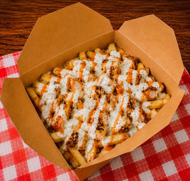 Fries topped with grilled chicken pieces, white sauce, and orange sauce in a cardboard box.