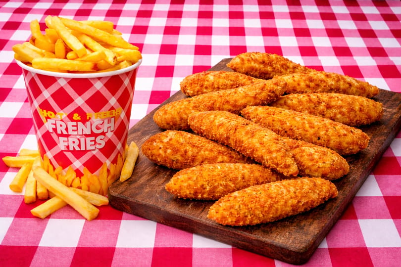 Crispy chicken tenders on a wooden board next to a cup of golden french fries on a red and white checkered tablecloth.