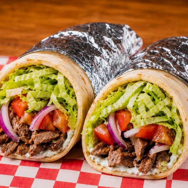 Two beef gyros with lettuce, tomatoes, and onions, partially wrapped in foil on a checkered paper.