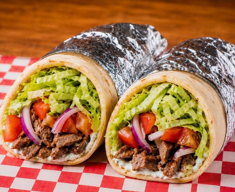 Two beef gyros with lettuce, tomatoes, and onions, partially wrapped in foil on a checkered paper.
