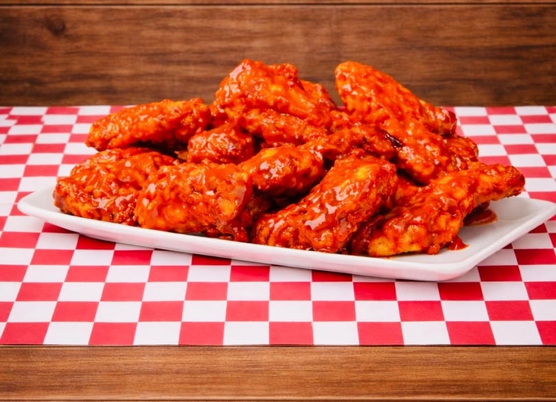 A white platter piled high with bright red, saucy chicken wings on a red and white checkered tablecloth.